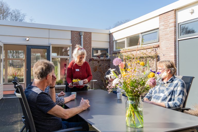 Twee personen aan tafel op een buitenplaats, een vrijwilliger brengt drinken
